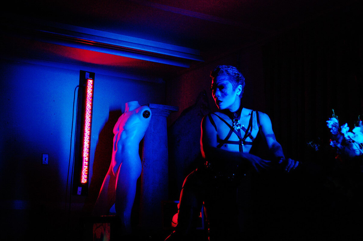 Maiden, wearing a harness and mounted on a chaise under bisexual lighting.